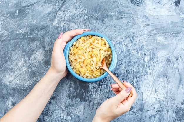 Bovenaanzicht geassorteerde pasta in kom en hand pasta mengen met houten lepel op grijze gips achtergrond. horizontaal