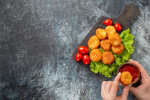 Bovenaanzicht gebakken kaasballetjes, cherrytomaatjes op snijplank, ketchupkom en kaasbal in vrouwelijke handen