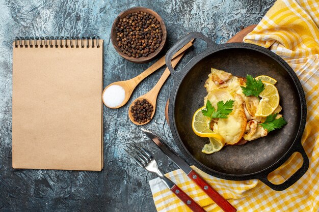 Bovenaanzicht gebakken vis in pan met citroen en peterselie kruiden in kom en houten lepels vork en mes notitieboekje op grijze achtergrond