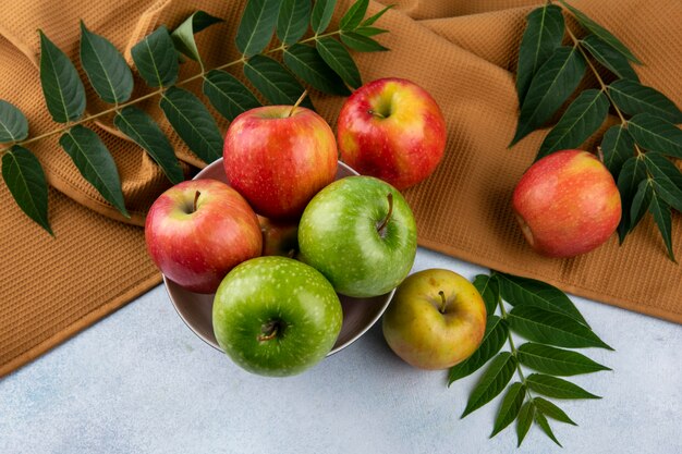 Bovenaanzicht gekleurde appels in een kom met bladtakken op een bruine handdoek op een grijze achtergrond