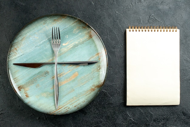 Bovenaanzicht gekruiste vork en mes op cyaan ronde plaat notebook op donkere tafel