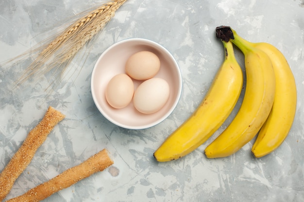 Bovenaanzicht gele bananen met rauwe eieren en broodje brood op licht wit bureau