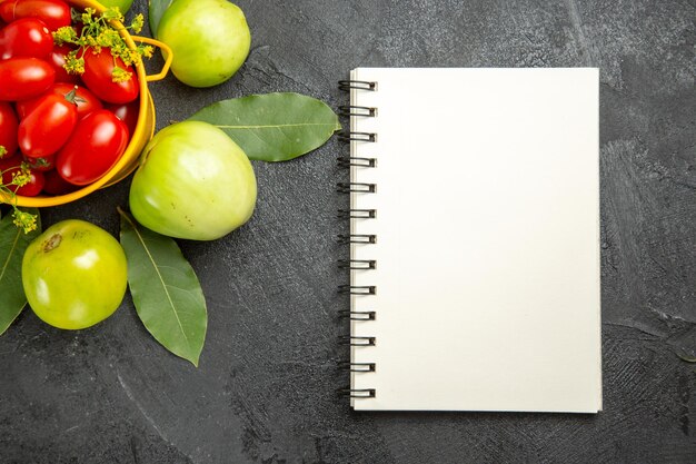 Bovenaanzicht gele emmer gevuld met kerstomaatjes en dille bloemen groene tomaten laurierblaadjes en een notitieboekje op donkere ondergrond