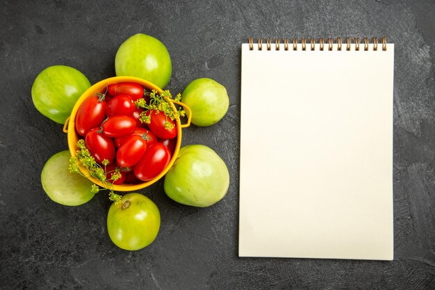 Bovenaanzicht gele emmer gevuld met kerstomaatjes en dillebloemen omringd met groene tomaten en een notitieboekje op donkere grond