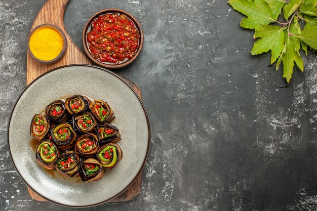 Bovenaanzicht gevulde auberginebroodjes in witte ovale plaat kurkuma in kom op houten serveerplank adjika op grijze ondergrond