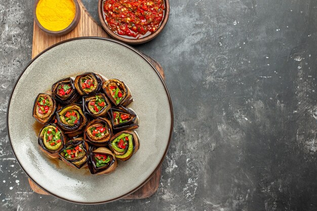 Bovenaanzicht gevulde auberginebroodjes in witte ovale plaat kurkuma in kom op houten serveerplank met handvat adjika op grijze achtergrond