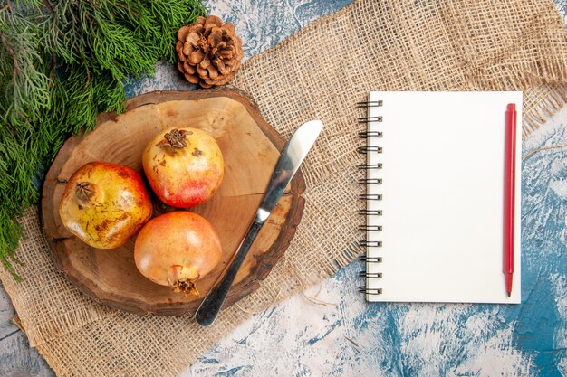 Bovenaanzicht granaatappels dinermes op ronde boom houten snijplank dennenboomtak notitieboekje met rode pen op blauw-wit oppervlak