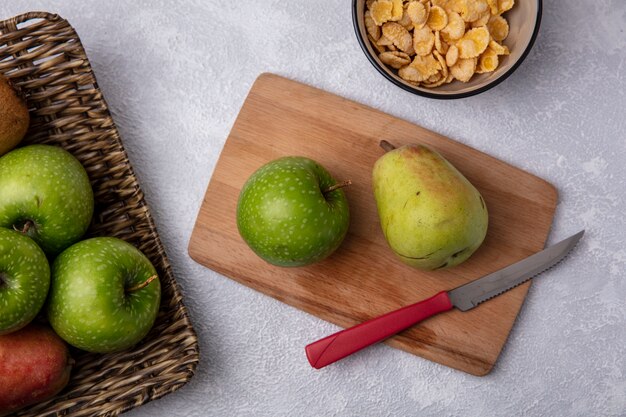 Bovenaanzicht groene appel met peer en mes op snijplank met cornflakes in kom op witte achtergrond