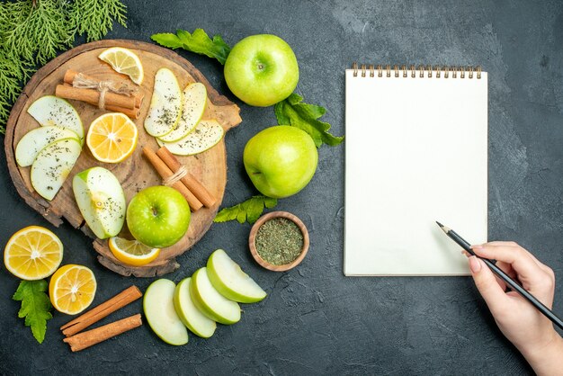 Bovenaanzicht groene appels kaneelstokjes en schijfjes citroen Appelschijfjes op een houten bord gesneden citroenen appels notebook potlood in vrouwelijke hand op zwarte tafel