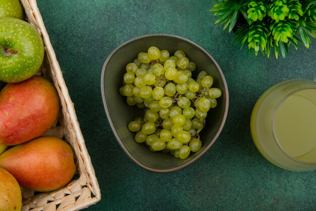 Bovenaanzicht groene druiven in een kom met een groene appel en peren in een mand met sap op een groene achtergrond
