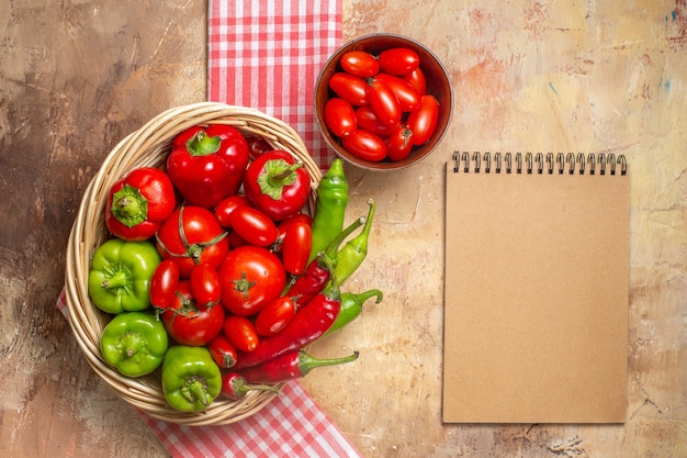 Bovenaanzicht groene en rode paprika's hete pepers tomaten in rieten mand kerstomaatjes in kom keukenhanddoek een notitieboekje op amberkleurige achtergrond