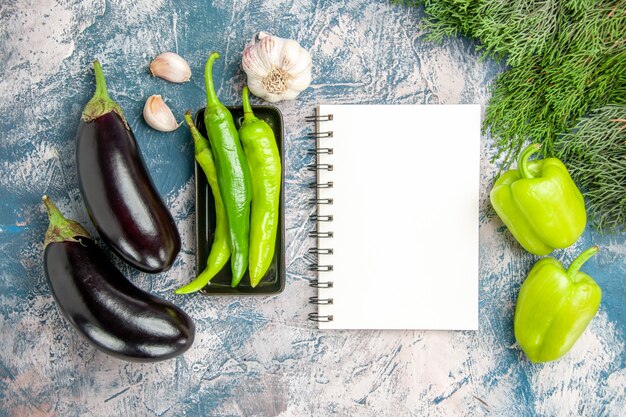 Bovenaanzicht groene hete pepers op zwarte plaat knoflook aubergines pepers een notitieboekje op blauw-witte achtergrond