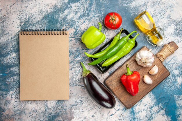 Bovenaanzicht groene hete pepers op zwarte plaat tomaat rode en groene paprika's knoflook op snijplank aubergine een notitieboekje op blauw-witte achtergrond