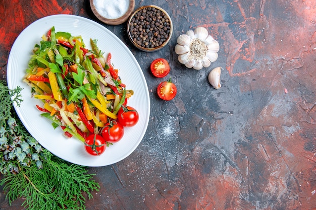 Bovenaanzicht groentesalade op ovale plaat cherry tomaten knoflook kruiden op donkerrode tafel vrije ruimte