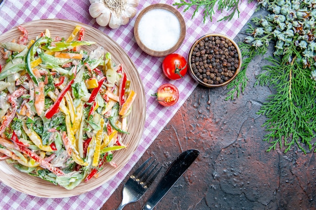 Bovenaanzicht groentesalade op plaat op tafelkleed vork en mes zout en zwarte peper dennen tak op donkerrode tafel