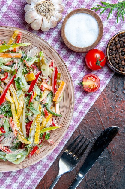 Bovenaanzicht groentesalade op plaat op tafelkleed vork en mes zout en zwarte peper knoflook op donkerrode tafel