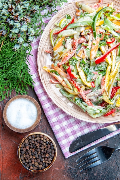 Bovenaanzicht groentesalade op plaat op tafelkleed vork en mes zout en zwarte peper pijnboomtakken op donkerrode tafel