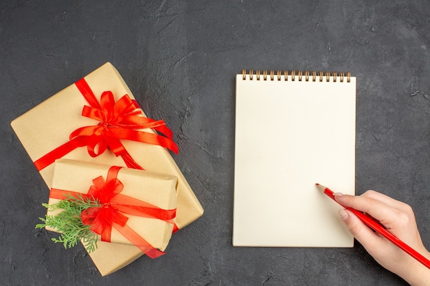 Bovenaanzicht grote en kleine kerstcadeaus in bruin papier gebonden met rood lint notebook potlood in vrouwelijke hand op donkere ondergrond