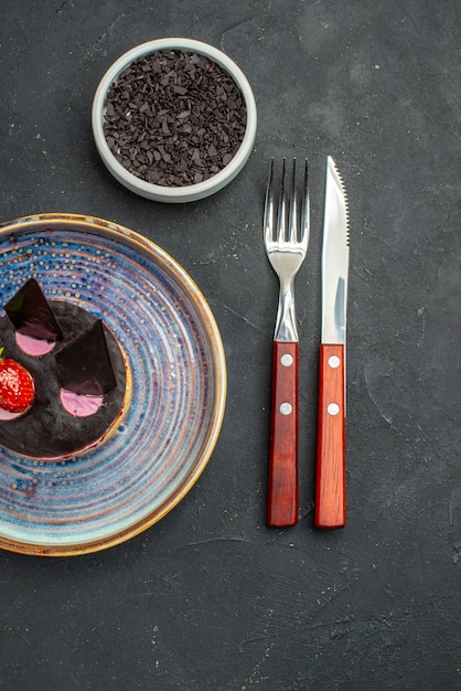 Bovenaanzicht heerlijke cheesecake met aardbei en chocolade op plaatkom met chocoladevork en mes op donkere geïsoleerde achtergrond