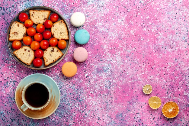 Bovenaanzicht heerlijke gesneden cake met zure verse pruimen macarons en kopje thee op het roze oppervlak