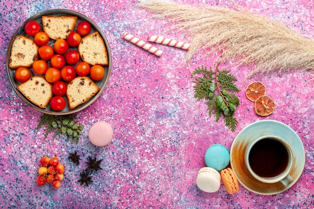 Bovenaanzicht heerlijke gesneden cake met zure verse pruimen macarons en thee op het roze oppervlak