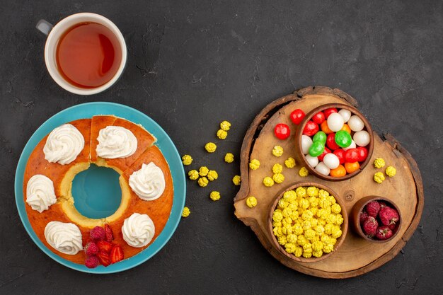 Bovenaanzicht heerlijke slagroomtaart met snoep en kopje thee op donkere achtergrond kandijsuiker cake koekjesdeeg fruit