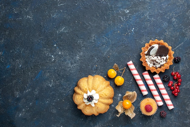 Bovenaanzicht in de verte van kleine heerlijke cake samen met koekje en roze stoksuikergoedvruchten op donker bureau, koektaartfruit