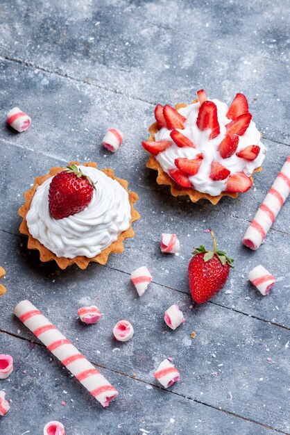 Bovenaanzicht in de verte van kleine romige cakes met gesneden en verse aardbeien, samen met stoksnoepjes op grijs, fruitcake zoet gebakken bessen