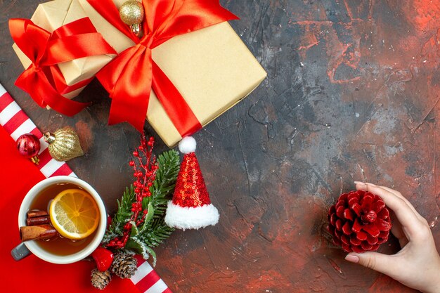 Bovenaanzicht kerstcadeaus gebonden met rood lint kerstmuts dennenappel in vrouwelijke hand op donkerrode tafel kopieerplaats