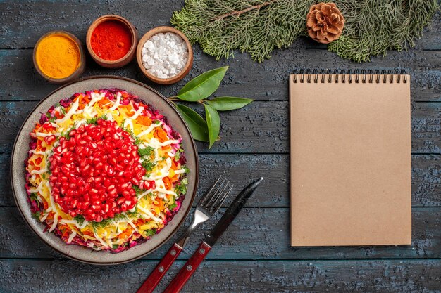 Bovenaanzicht kerstschotel met granaatappel wortelen aardappelen naast de crème notebook kommen van verschillende kruiden vuren takken met kegels op tafel