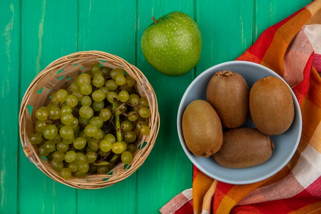 Bovenaanzicht kiwi in een kom met druiven in een mand en een groene appel op een geruite handdoek op een groene achtergrond