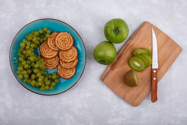 Bovenaanzicht kiwi plakjes met een mes op een snijplank met groene appels en groene druiven met koekjes op een plaat op een witte achtergrond