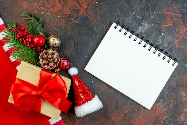 Bovenaanzicht klein cadeautje vastgebonden met rood lint xmas boomtak mini kerstmuts notitieblok op donkerrode tafel