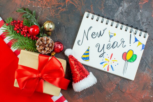 Bovenaanzicht klein cadeautje vastgebonden met rood lint xmas boomtak nieuwjaar geschreven op notitieblok op donkerrode tafel