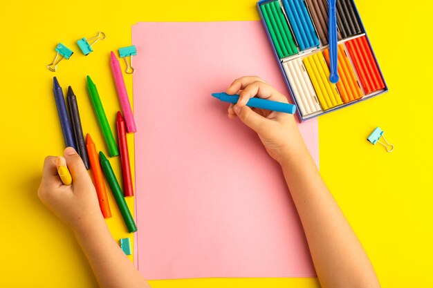 Bovenaanzicht klein kind met kleurrijke potloden op roze papier op geel oppervlak