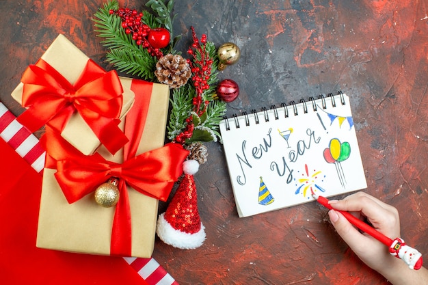 Bovenaanzicht kleine geschenken gebonden met rood lint kerstmuts xmas boomtak nieuwjaar geschreven op kladblok pen in vrouwelijke hand op donkerrode tafel