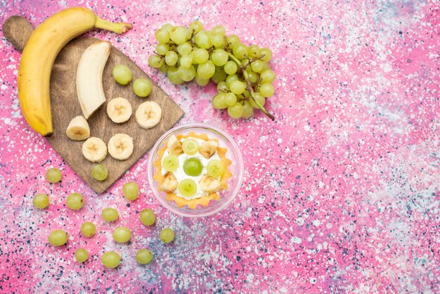 Bovenaanzicht kleine heerlijke cake met room en gesneden bananen en druiven op het heldere oppervlak fruit zoet