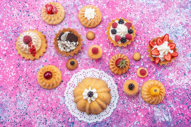 Bovenaanzicht kleine lekkere cakes met room samen met verschillende bessen op de licht-heldere tafel cake biscuit bes zoete bak thee