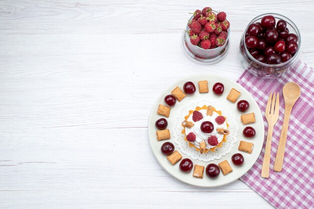 Bovenaanzicht kleine romige cake met frambozen en kleine koekjes samen met kersen op de lichttafel fruitcake zoete bessenroom kers