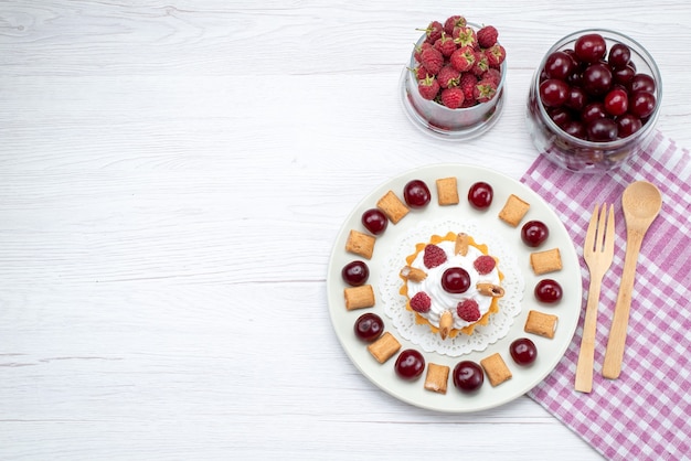 Bovenaanzicht kleine romige cake met frambozen en kleine koekjes samen met kersen op de lichttafel fruitcake zoete bessenroom kers