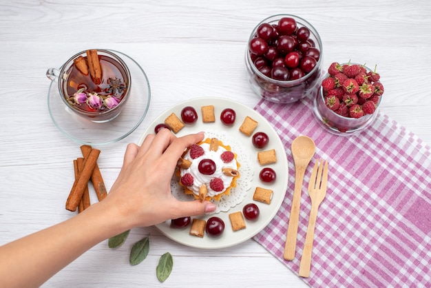 Bovenaanzicht kleine romige cake met frambozen, kersen en kleine koekjes thee kaneel op de lichttafel fruitcake bessenroom suiker thee