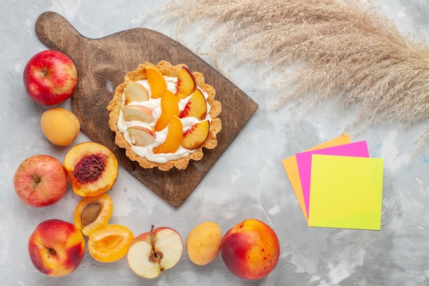 Bovenaanzicht kleine romige cake met gesneden fruit en witte room samen met vers fruit op licht wit bureau fruitcake biscuit zoete room bakken