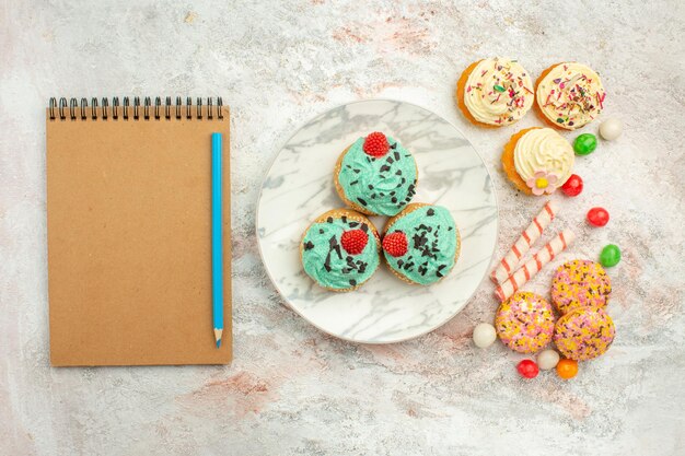 Bovenaanzicht kleine slagroomtaarten met kleurrijke snoepjes en koekjes op wit oppervlak goodie regenboog snoep dessert kleur cake