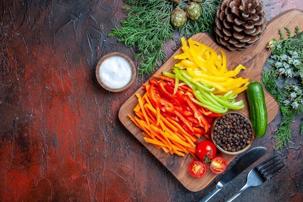 Bovenaanzicht kleurrijke gesneden paprika zwarte peper tomaten komkommer op snijplank zout vork en mes op donkerrode tafel met vrije ruimte