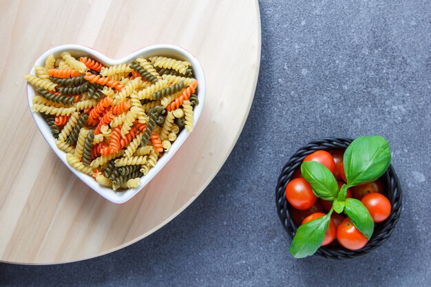 Bovenaanzicht kleurrijke macaroni pasta in hartvormige kom met tomaten, bladeren, op houten platform en grijze ondergrond. horizontaal