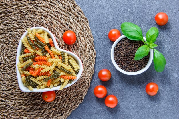 Bovenaanzicht kleurrijke macaroni pasta in hartvormige kom met tomaten, zwarte peper op onderzetter en grijze ondergrond. horizontaal