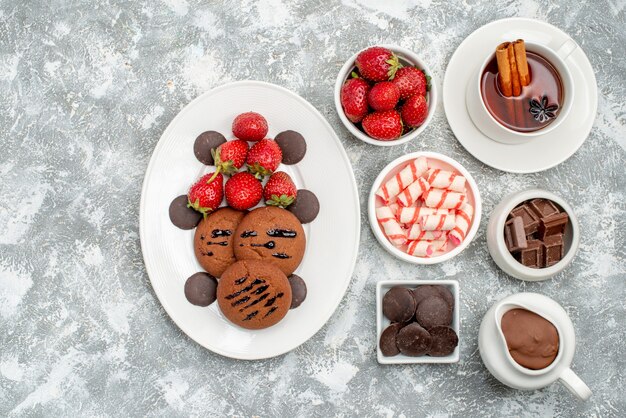 Bovenaanzicht koekjes aardbeien en chocoladeschaaltjes met cacao snoep aardbeien chocolaatjes en thee met kaneel aan de rechterkant van de grijs-witte tafel