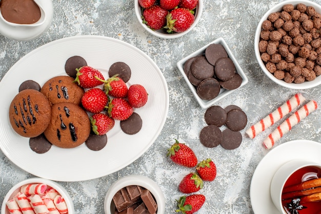 Bovenaanzicht koekjes aardbeien en ronde chocolaatjes op de ovale plaat schalen met snoepjes aardbeien chocolaatjes granen cacao en kaneelthee op de grijs-witte tafel