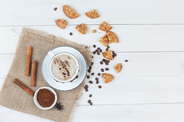 Bovenaanzicht koffie in beker met koffiebonen, gemalen koffie, koekjes, kaneelstokjes op houten en stuk zak achtergrond. horizontaal