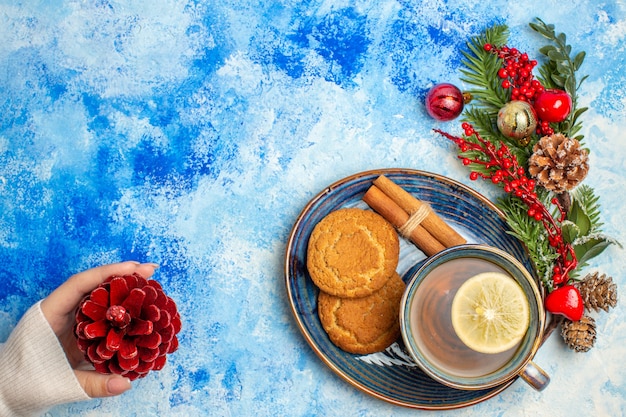 Bovenaanzicht kopje thee citroen schijfjes kaneelstokjes koekjes op schotel rode dennenappel in vrouwelijke hand op blauwe tafel vrije plaats